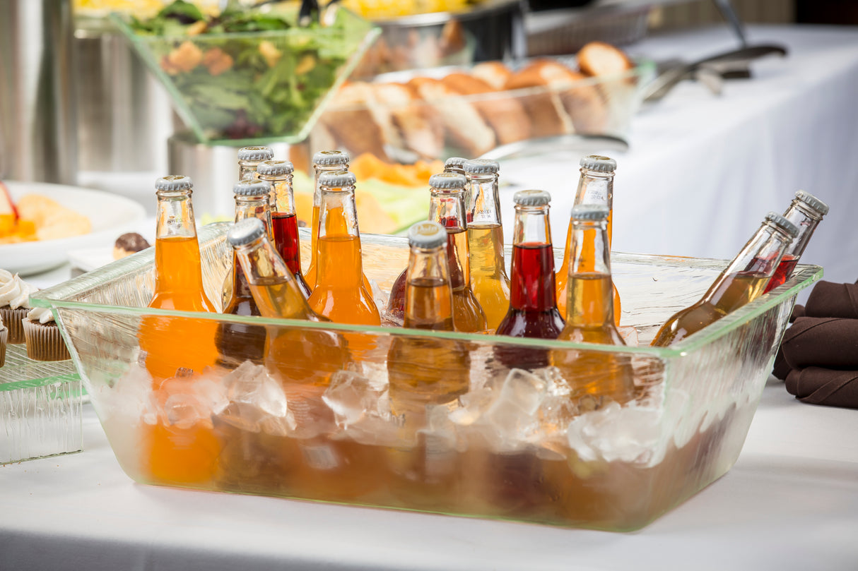 An acrylic beverage tub filled with ice holds various bottled soft drinks in vibrant colors. The dishwasher safe container sits on a table covered with a white tablecloth, with blurred food trays and bread loaves in the background.