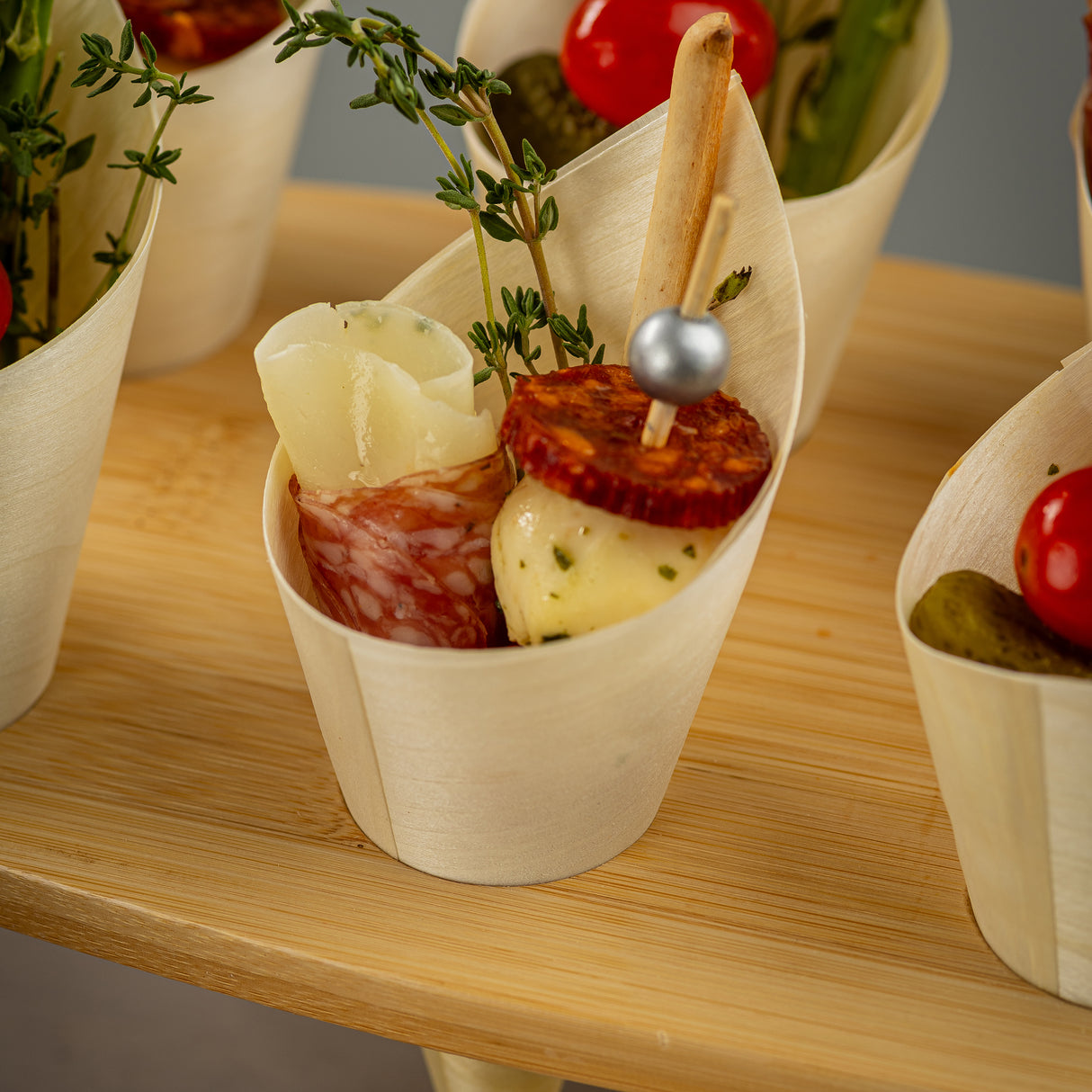An eco-friendly disposable mini serving cone made from biodegradable pinewood holds rolled salami, a cheese curl, a skewer with herb-coated cheese and chorizo, a breadstick, cherry tomato, thyme sprigs, and a pickle—all arranged neatly on a wooden board.