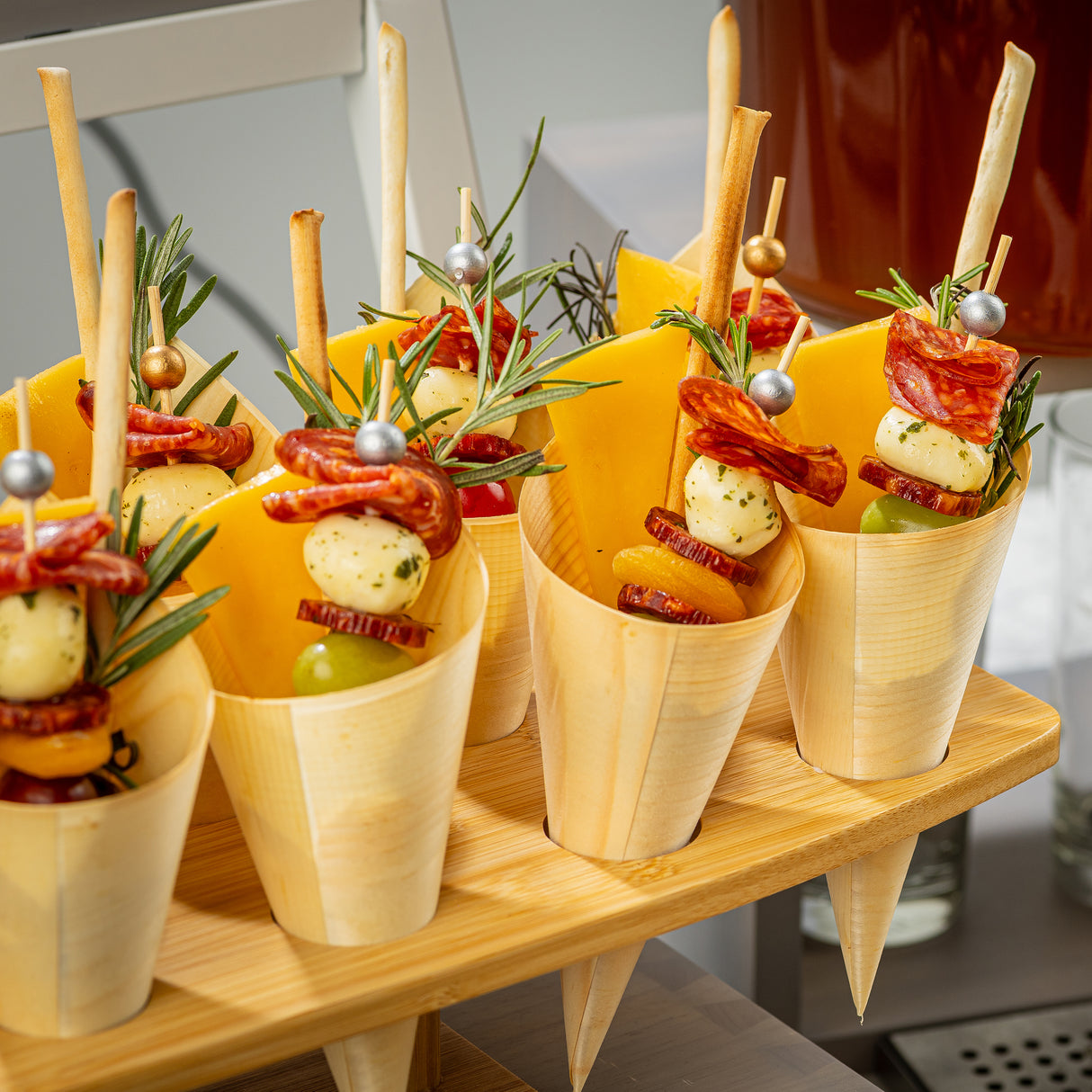 Wooden cones, made from biodegradable, eco-friendly materials, are filled with cheese slices, rolled salami, mozzarella balls, grapes, breadsticks, and rosemary. The disposable serving cones are artfully arranged on a wooden tray for easy appetizers.