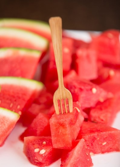 Close-up of a TableCraft bamboo fork stuck into a cube of fresh, vibrant red watermelon. In the background, more watermelon cubes and slices with green rind evoke a refreshing, summery fruit platter. Seeds are visible in some cubes.