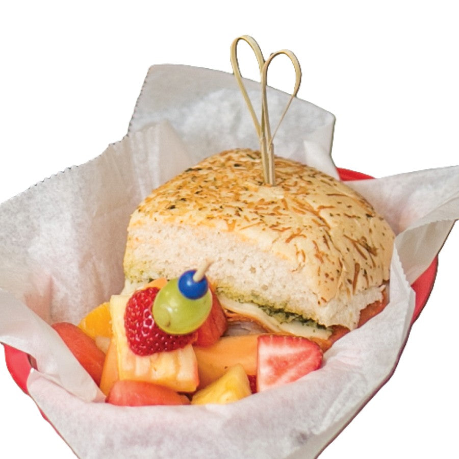 A sandwich with leafy greens and deli meat on herbed focaccia bread, secured with a Heart Looped Pick by TableCraft, sits in a red basket lined with white parchment. Beside it is a fruit kebab with green grape, strawberry, cheese, and various cut fruits.