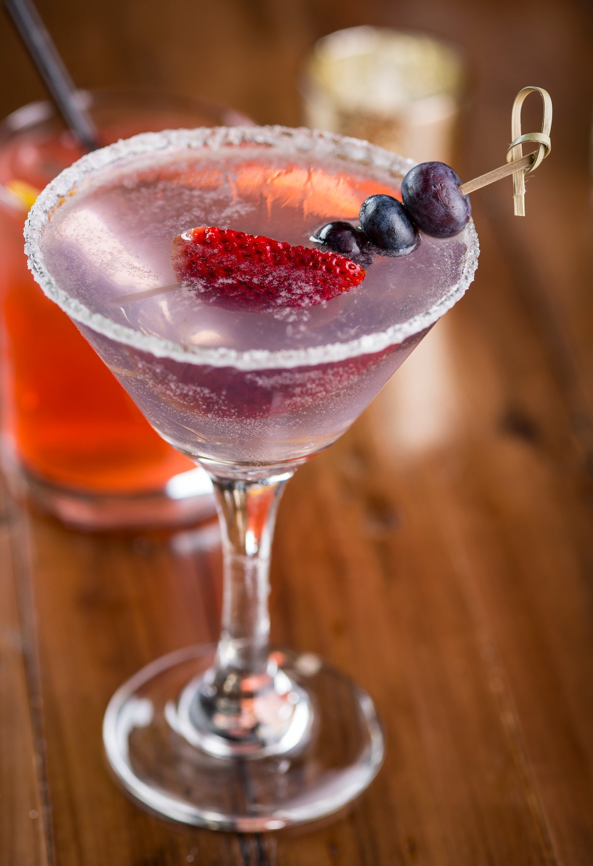 A purple cocktail in a martini glass with a sugared rim, garnished with fresh fruit on a TableCraft Knot Pick. The glass sits on a wooden table, with a blurry orange drink in the background.