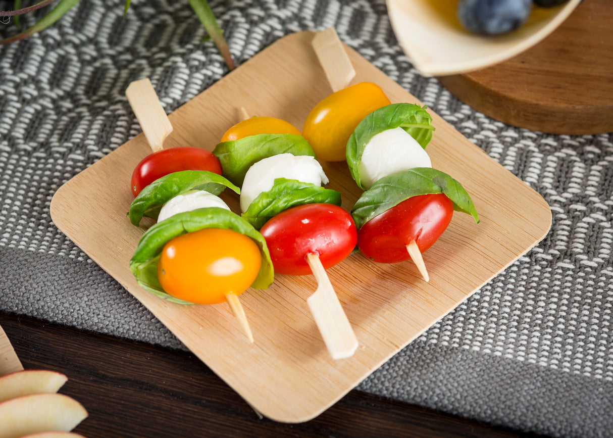 Three TableCraft Paddle Pick skewers on a square wooden plate hold yellow and red grape tomatoes, fresh basil leaves, and small mozzarella balls. The background features a gray woven placemat and part of a wooden table.
