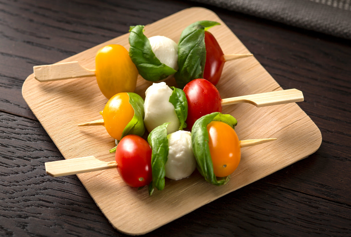 Two bamboo Paddle Picks by TableCraft rest on a square wooden plate, each threaded with cherry tomatoes, fresh basil leaves, and mozzarella balls, arranged in alternating colors for a fresh and colorful appetizer display on a dark table.