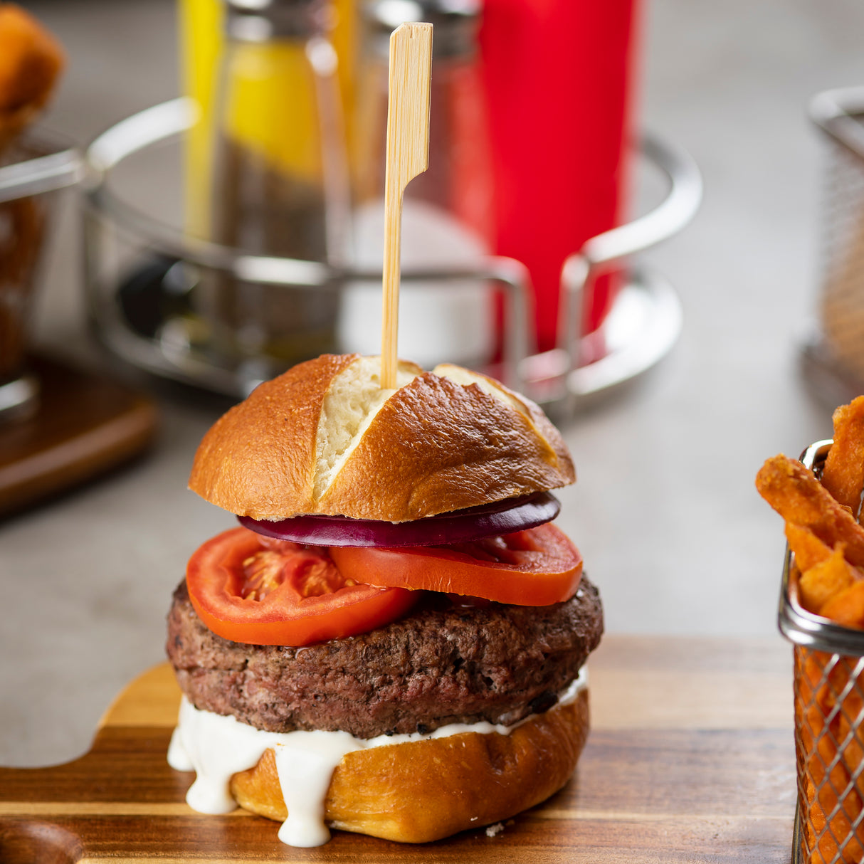 A juicy beef burger with sliced tomato, red onion, and white sauce sits on a wooden board. The burger is held together with a TableCraft bamboo paddle pick on a shiny brioche bun. Sweet potato fries are in a metal basket; condiments blur in the background.