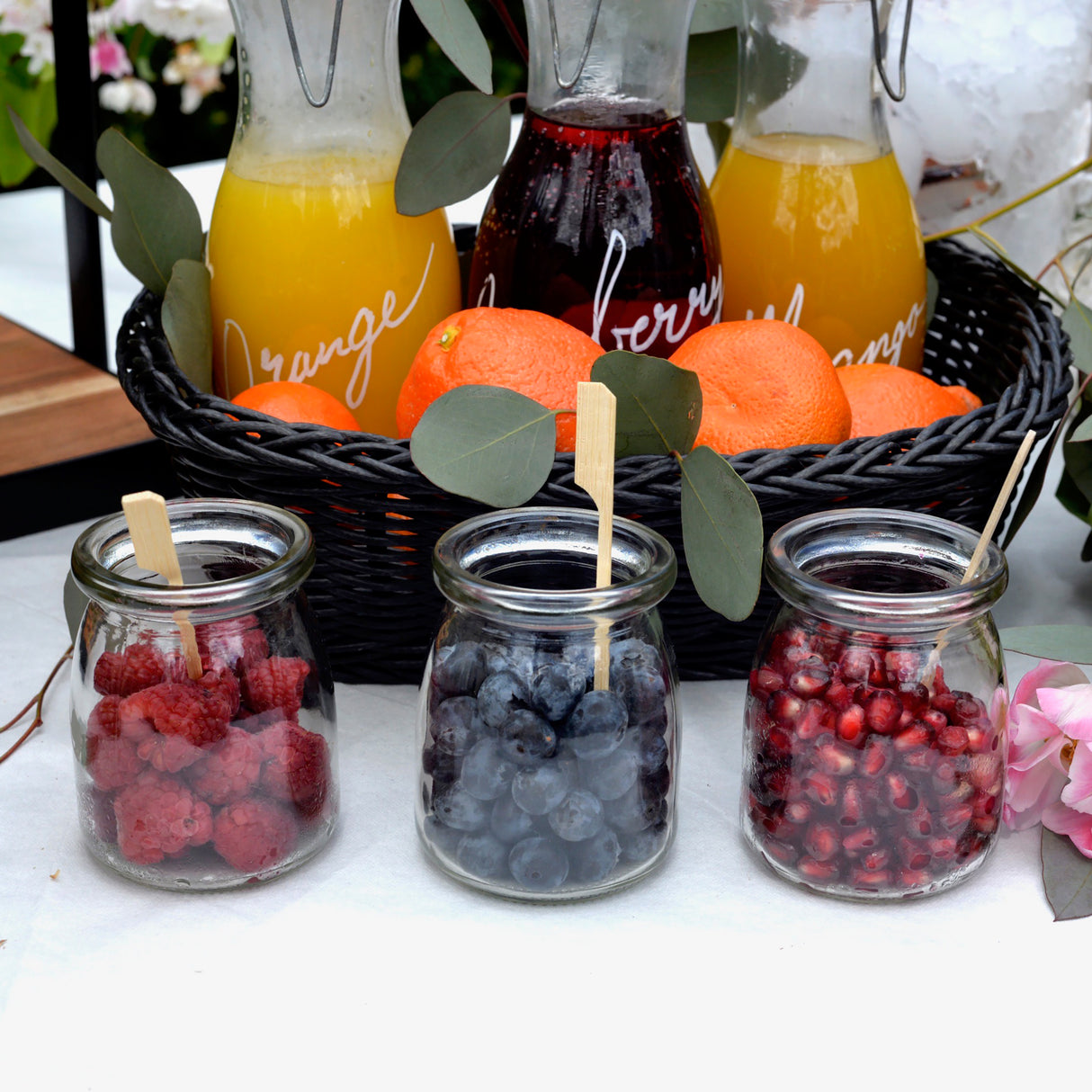 Three glass jars filled with raspberries, blueberries, and pomegranate seeds are in front of a black basket holding oranges and juice pitchers. TableCraft Products like a bamboo paddle pick add flair to this vibrant scene decorated with green leaves and a pink flower.