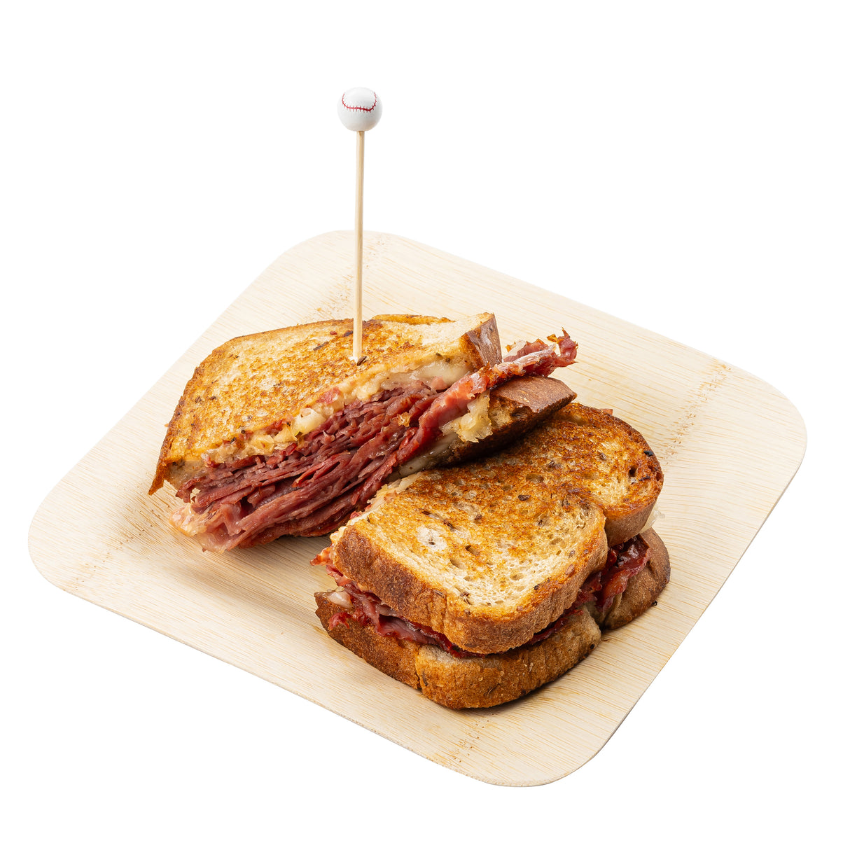A Reuben sandwich cut in half, stacked on a light wooden plate. Layers of corned beef, melted cheese, sauerkraut, and dressing fill toasted rye bread. A decorative bamboo skewer with a round top holds the halves together.