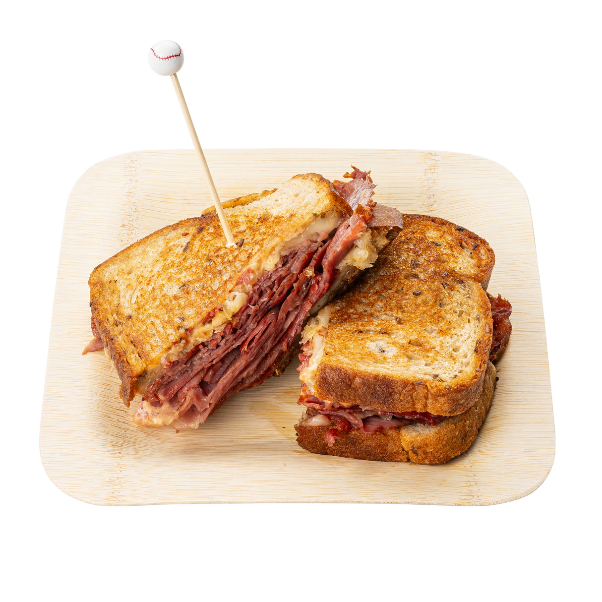 A Reuben sandwich cut in half sits on a wooden plate. Layers of corned beef, sauerkraut, and melted cheese fill toasted rye bread. One half is secured with a Sport Pick topped with a small baseball for a playful finish.