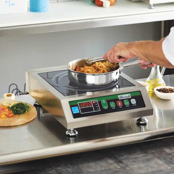 A person is cooking pasta in a metal pan on a Commercial Induction Cooker with digital controls. Chopped vegetables, herbs, and a bottle of oil are on the counter nearby, suggesting meal preparation in a commercial kitchen setting.