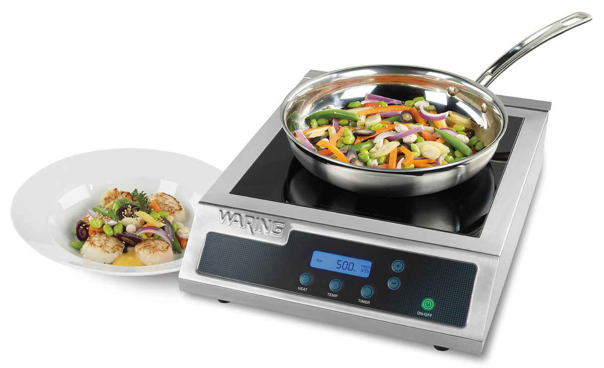 A stainless steel induction range single hob cooktop with a digital display shows a frying pan of colorful stir-fried vegetables. Beside it, on the countertop, is a white plate with gourmet seafood and vegetable dish, garnished with herbs.