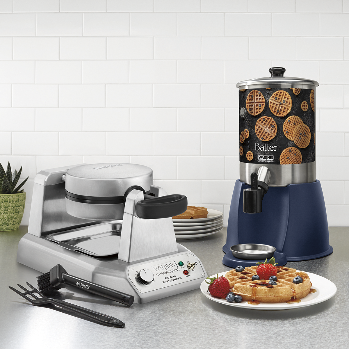 A stainless steel Belgian waffle maker with a waffle inside sits next to a 2 gallon batter dispenser labeled with a waffle image. Plates of golden waffles topped with strawberries, blueberries, and syrup sit in front. A fork, knife, and plant are also visible on the counter.