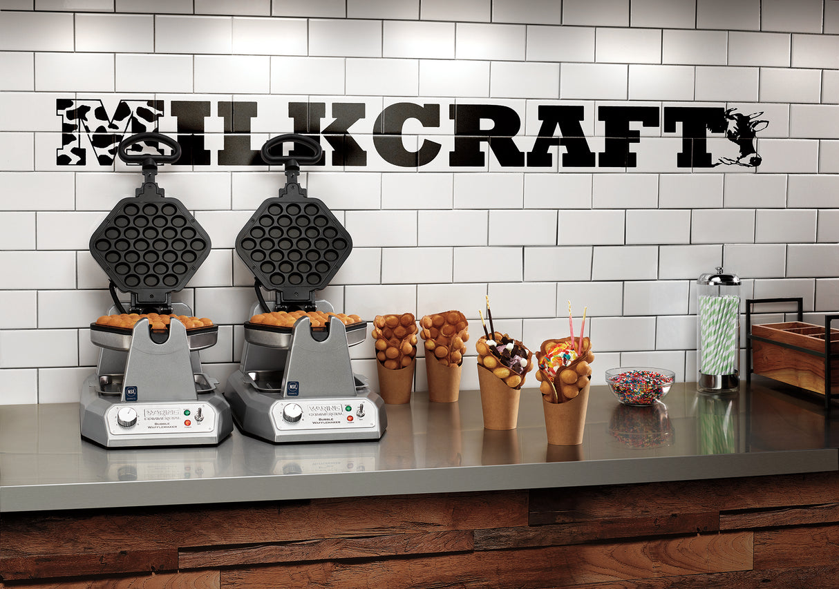 Two Waring bubble waffle makers with non-stick plates sit on a counter beneath a MILKCRAFT sign. In front, bubble waffles in cones are topped with ice cream and sprinkles, with green-striped straws and a bowl of sprinkles nearby.