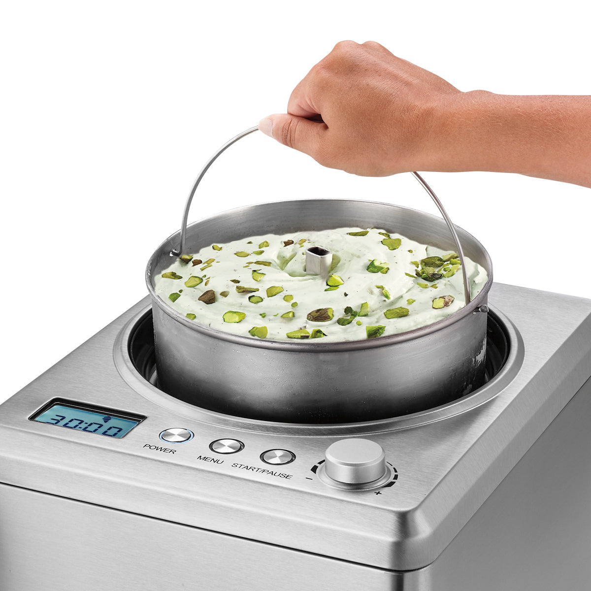 A hand lifts a metal container filled with pistachio-topped ice cream from an Ice Cream Maker with a built-in compressor. The silver machine features a digital display reading 30:00, several buttons, and a convenient control dial.