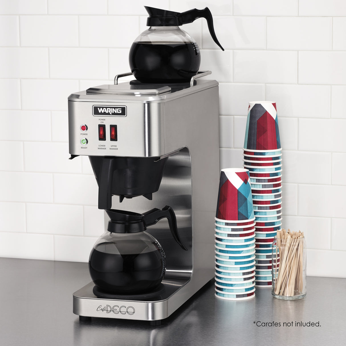 A stainless steel pourover coffee brewer with two glass carafes, one on a self regulating warmer below and one above, sits on a countertop. Nearby are stacked geometric-patterned paper cups and a container of wooden stir sticks. White tile backsplash is behind.