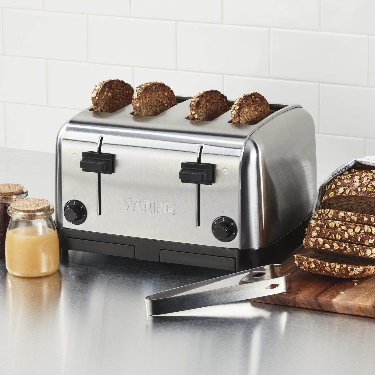 A stainless steel 4 slice toaster with extra wide slots toasts multigrain bread. Nearby are a jar of honey, jam, metal tongs, and a wooden board with sliced multigrain bread on a clean counter against white subway tiles.