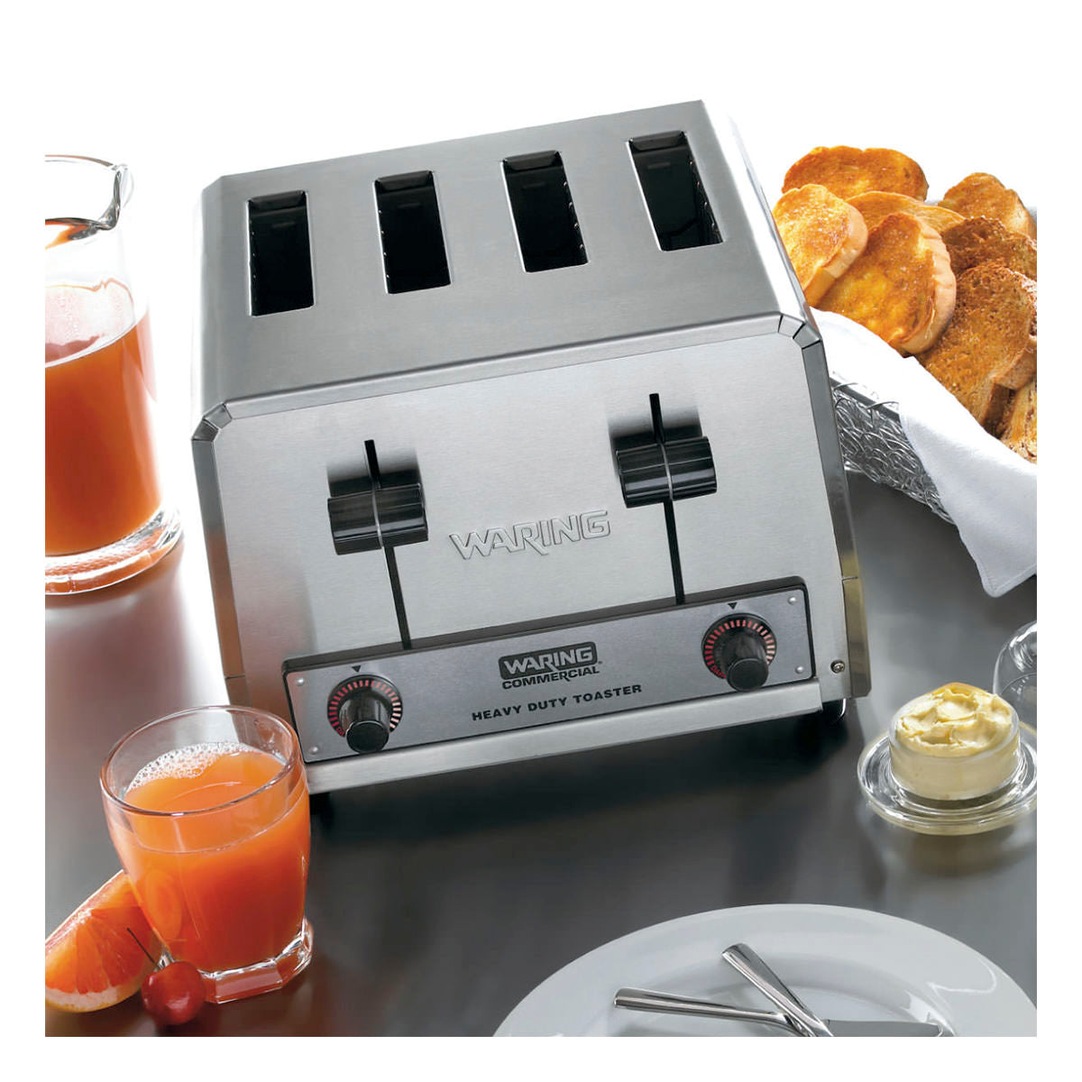 A stainless steel 4 slice toaster sits on a table with its controls visible. Surrounding it are a glass of orange juice, a plate with toast, a bowl of butter, an orange slice, and a basket of toasted bread. The table is set with utensils.