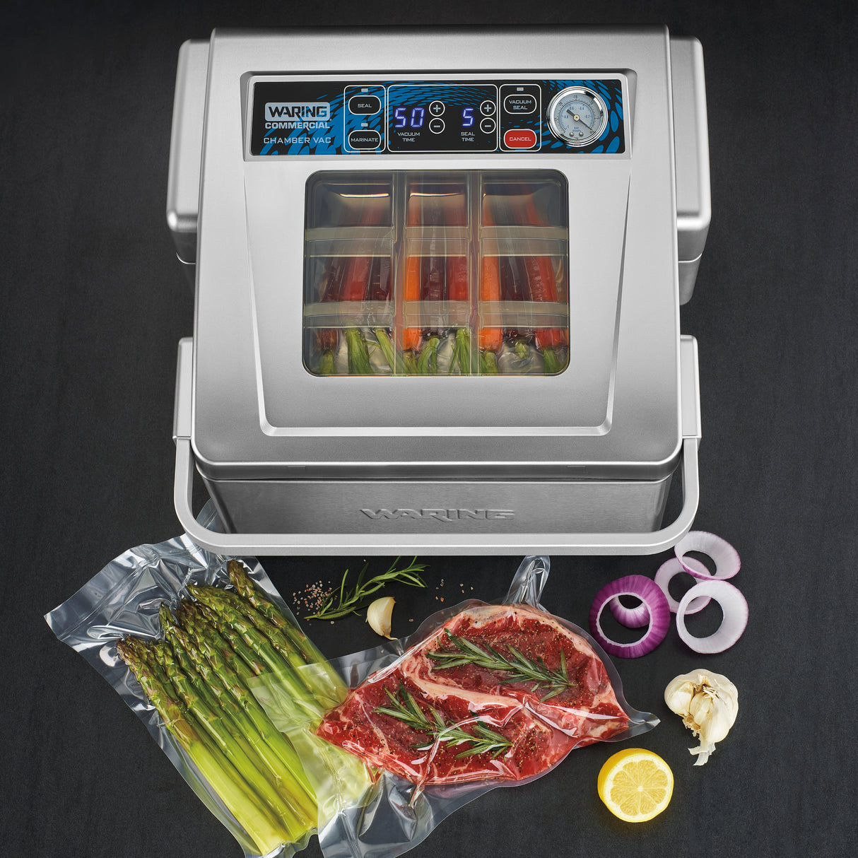 A Waring vacuum packaging machine sits on a dark countertop with its lid open, revealing bags of vegetables inside. In front are vacuum-sealed asparagus and steak with herbs, plus cut onions, garlic, rosemary, pepper, and sliced lemon for garnish.