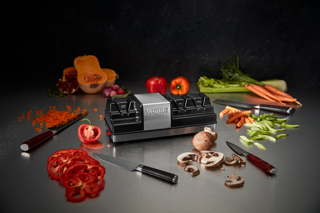 A kitchen scene centers on an electric knife sharpener, surrounded by sliced and whole tomatoes, peppers, carrots, mushrooms, celery, and squash. Three knives rest on a reflective countertop, with the sharpener’s grinding wheels subtly in view.