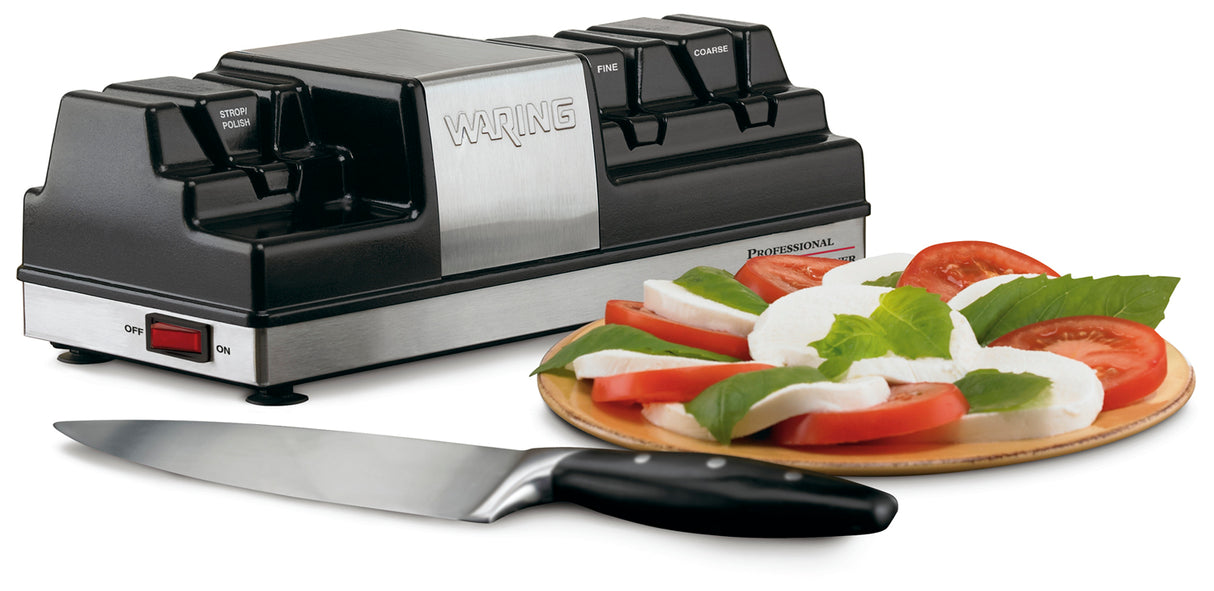 A Waring electric knife sharpener with grinding wheels sits on a white surface next to a plate of sliced tomatoes, mozzarella, and basil. A sharp kitchen knife in front highlights the electric sharpener’s role in effortless food prep.