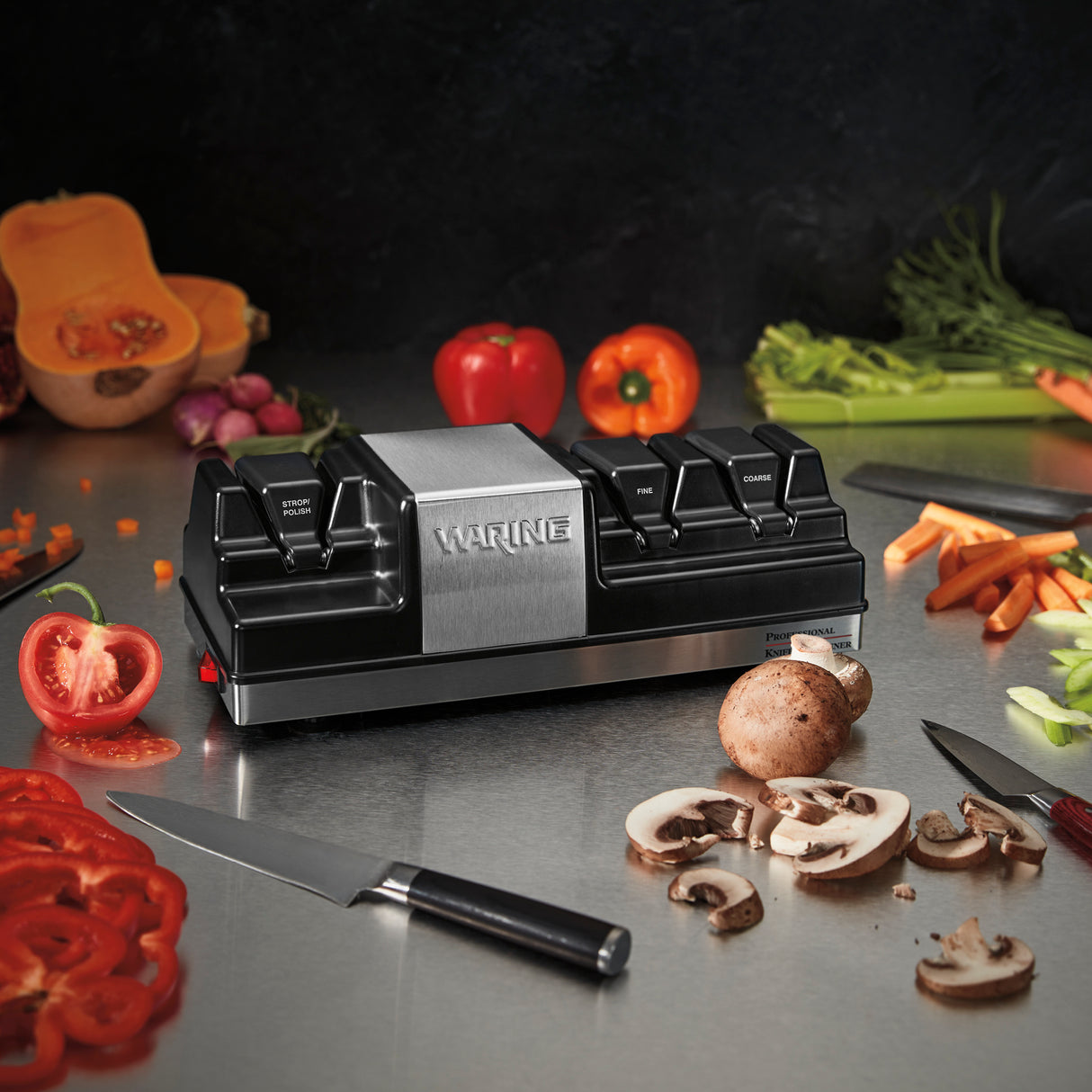 A Waring electric Knife Sharpener with powerful grinding wheels sits on a stainless steel countertop, surrounded by sliced tomatoes, mushrooms, carrots, and bell peppers. A chef’s knife lies nearby, with fresh vegetables and a dark backsplash in the background.