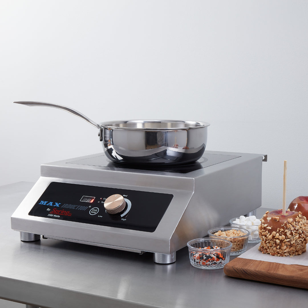 A stainless steel saucepan sits on a MAX Induction Cook & Hold Range countertop induction cooktop. Nearby are a candy apple on a wooden board, a glass bowl of mini marshmallows, and two small bowls with chopped nuts and colorful sprinkles.