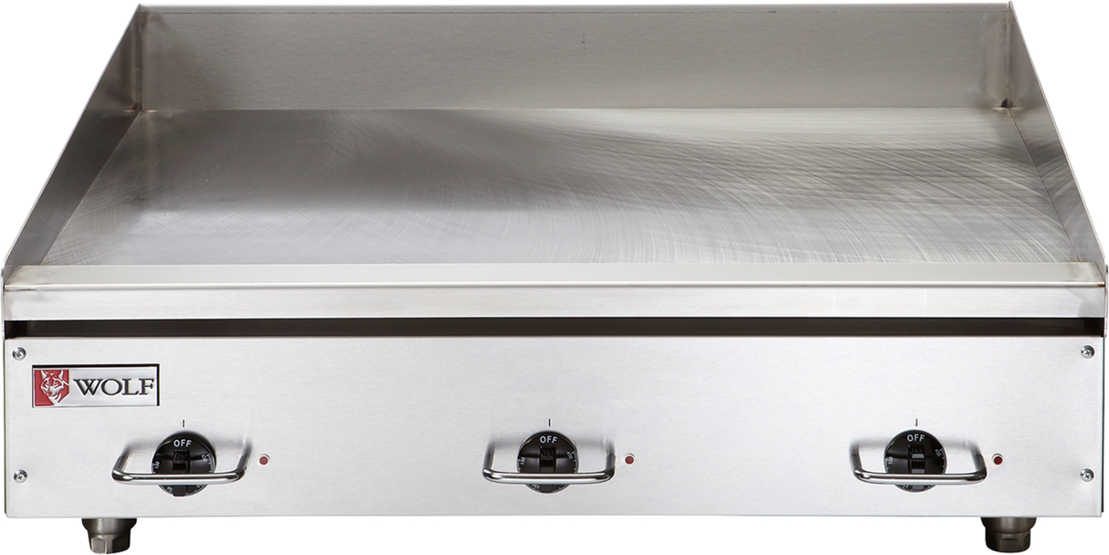 A stainless steel commercial electric griddle with a smooth flat cooking surface, three control knobs at the front, and a raised back and side splash guard. The Wolf brand logo is visible on the front left corner of this countertop cooking appliance.