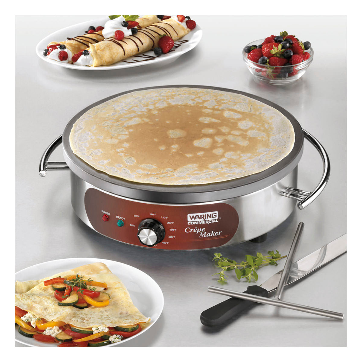 A Waring Commercial crepe maker with a cast iron cook surface and adjustable thermostat sits on a metal counter. Plates of rolled crepes topped with chocolate and berries, a bowl of mixed berries, and utensils surround it.