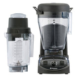 Two clear Vitamix XL Blender containers with black lids are shown, one upright on a commercial blender base with control knobs, the other resting on its side. Both containers feature measurement markings and metal blades visible at the bottom.
