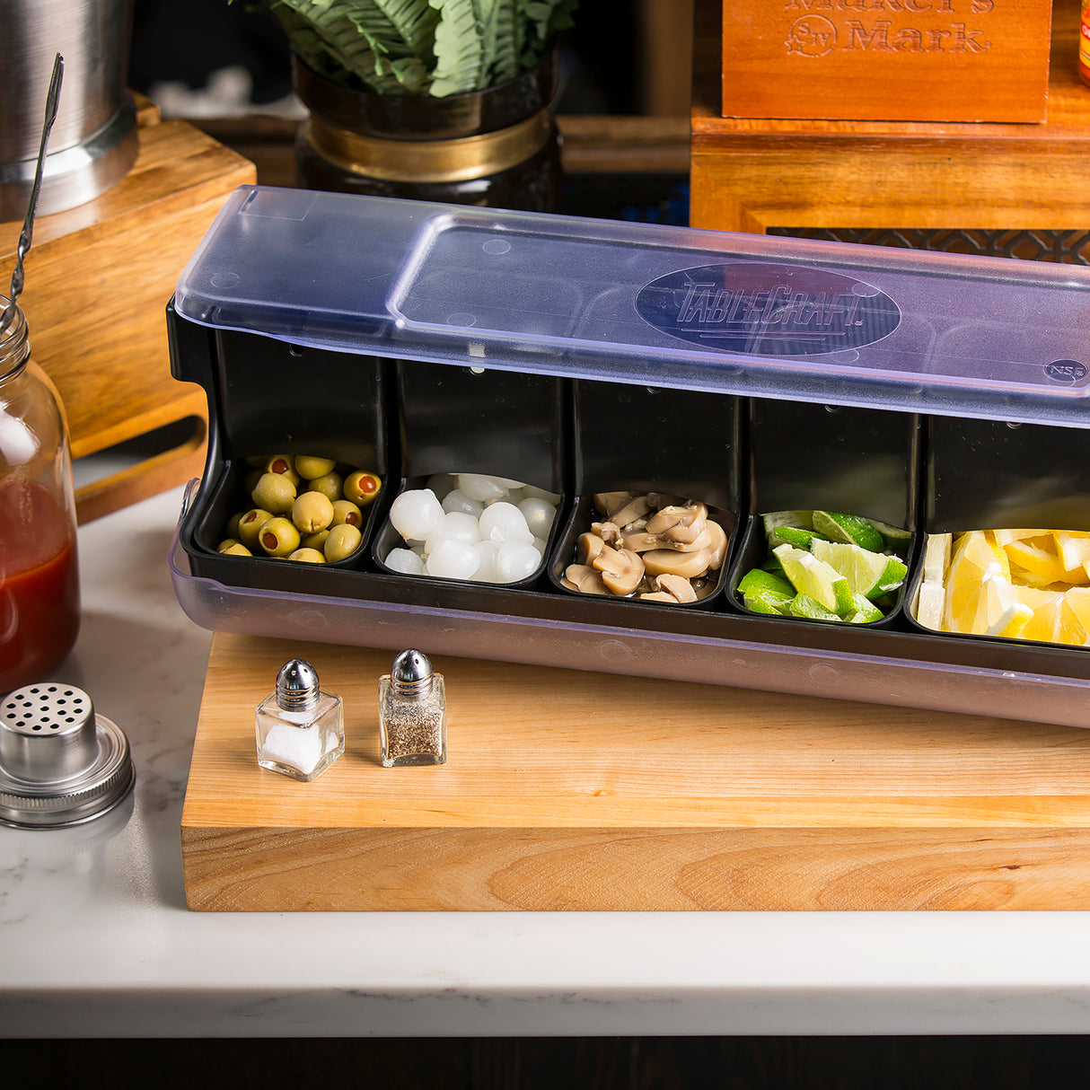 A black bar garnish tray with five compartments—dishwasher safe for easy cleaning—holds green olives, cocktail onions, mushrooms, lime wedges, and lemon wedges on a wooden board. Nearby are salt and pepper shakers, a jar of red liquid, and assorted bar tools.