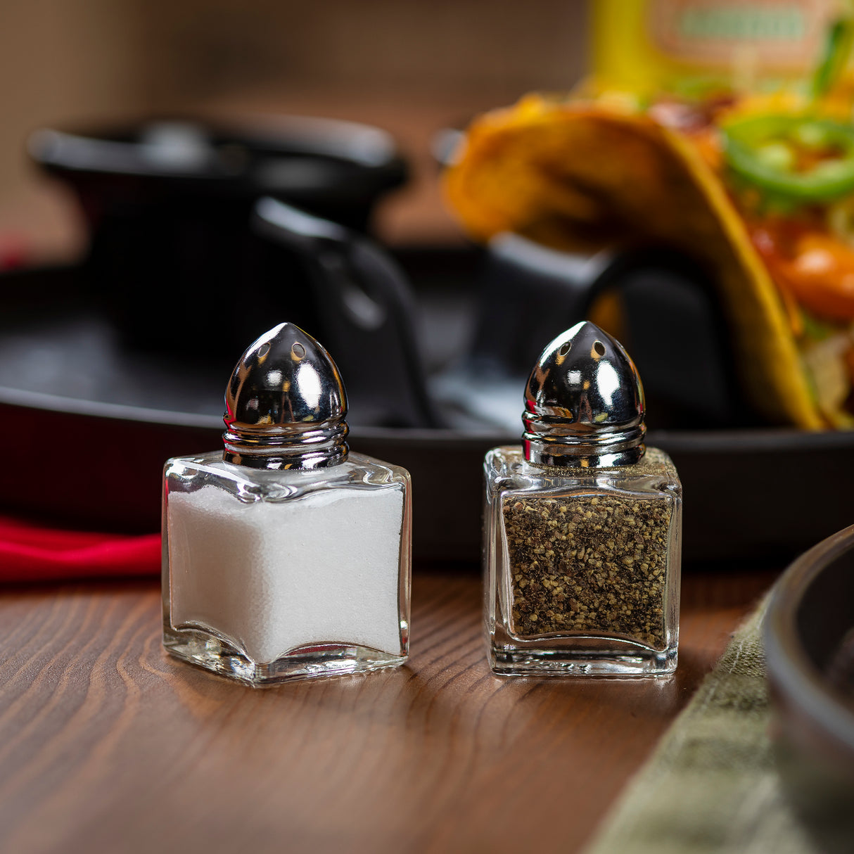 Two cube glass Salt/Pepper Shakers with silver tops sit on a wooden table; the left holds salt and the right pepper. Both are dishwasher safe. In the background, a taco on a black plate is partially visible, slightly out of focus.