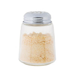 A modern glass cheese shaker with a shiny metal lid, filled with finely ground powder resembling parmesan cheese, stands out against a plain white background. This stylish piece is also dishwasher safe for easy cleaning.