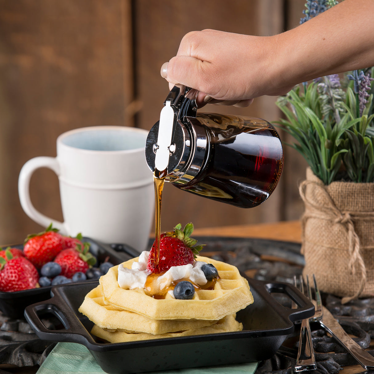 A hand uses a TableCraft replacement syrup dispenser top with a chrome plated finish to pour syrup onto waffles topped with whipped cream, strawberries, and blueberries in a black dish. Fresh fruit and potted plants decorate the rustic table nearby.