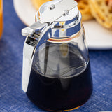 A 12 oz Modern Glass Dispenser with a chrome plated top sits on a blue tablecloth, filled with dark syrup. Waffles on a white plate are blurred in the background.