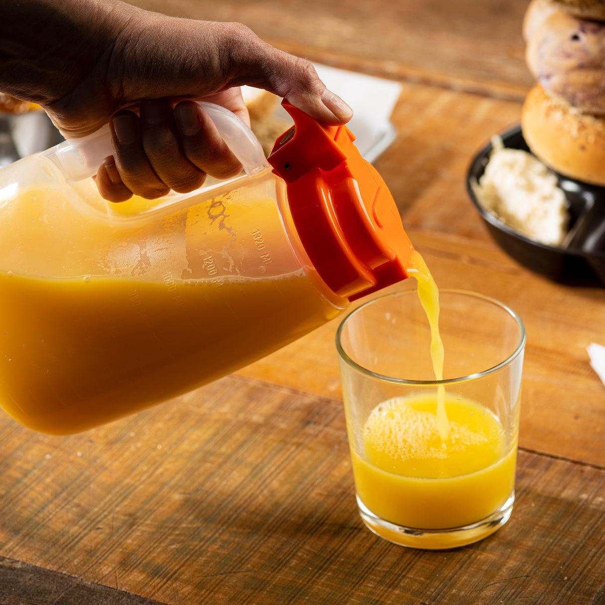A hand pours orange juice from a large, dishwasher safe TableCraft plastic pitcher with an orange lid into a clear glass on a wooden table. In the background, a black bowl holds a bagel and cream cheese, creating a cozy breakfast scene.
