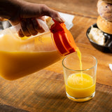 A hand pours orange juice from a large, dishwasher safe TableCraft plastic pitcher with an orange lid into a clear glass on a wooden table. In the background, a black bowl holds a bagel and cream cheese, creating a cozy breakfast scene.
