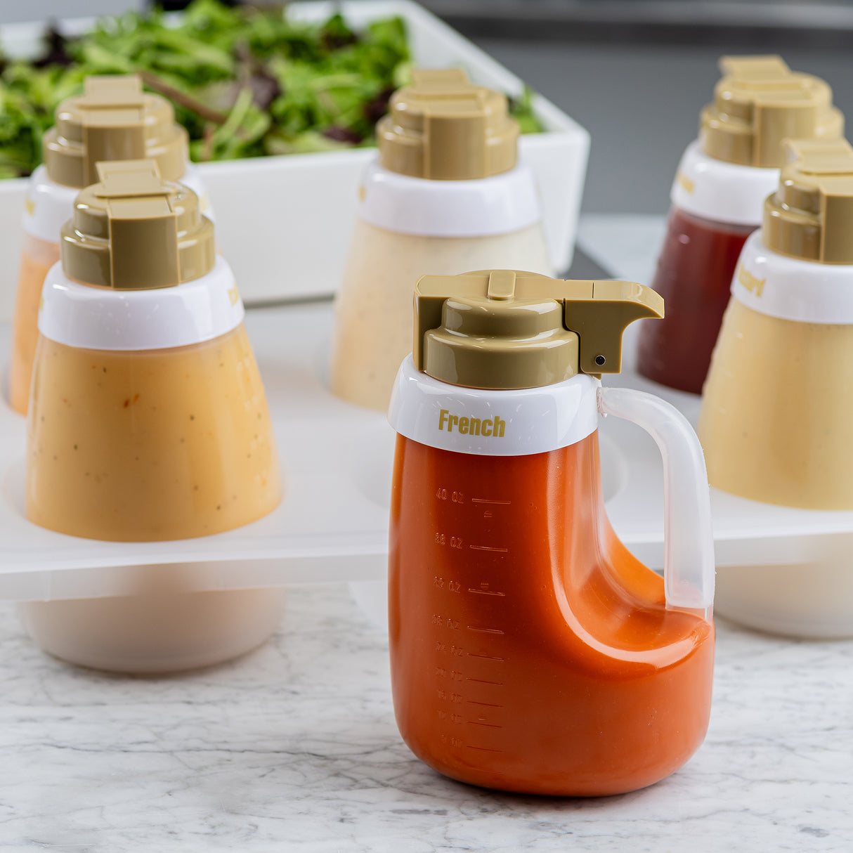 Several pump-top condiment bottles filled with different salad dressings are arranged on a white plastic tray beside a bowl of mixed greens. The Salad Dressing Dispenser Set, with dishwasher safe bottles, features the front bottle labeled “French.”.