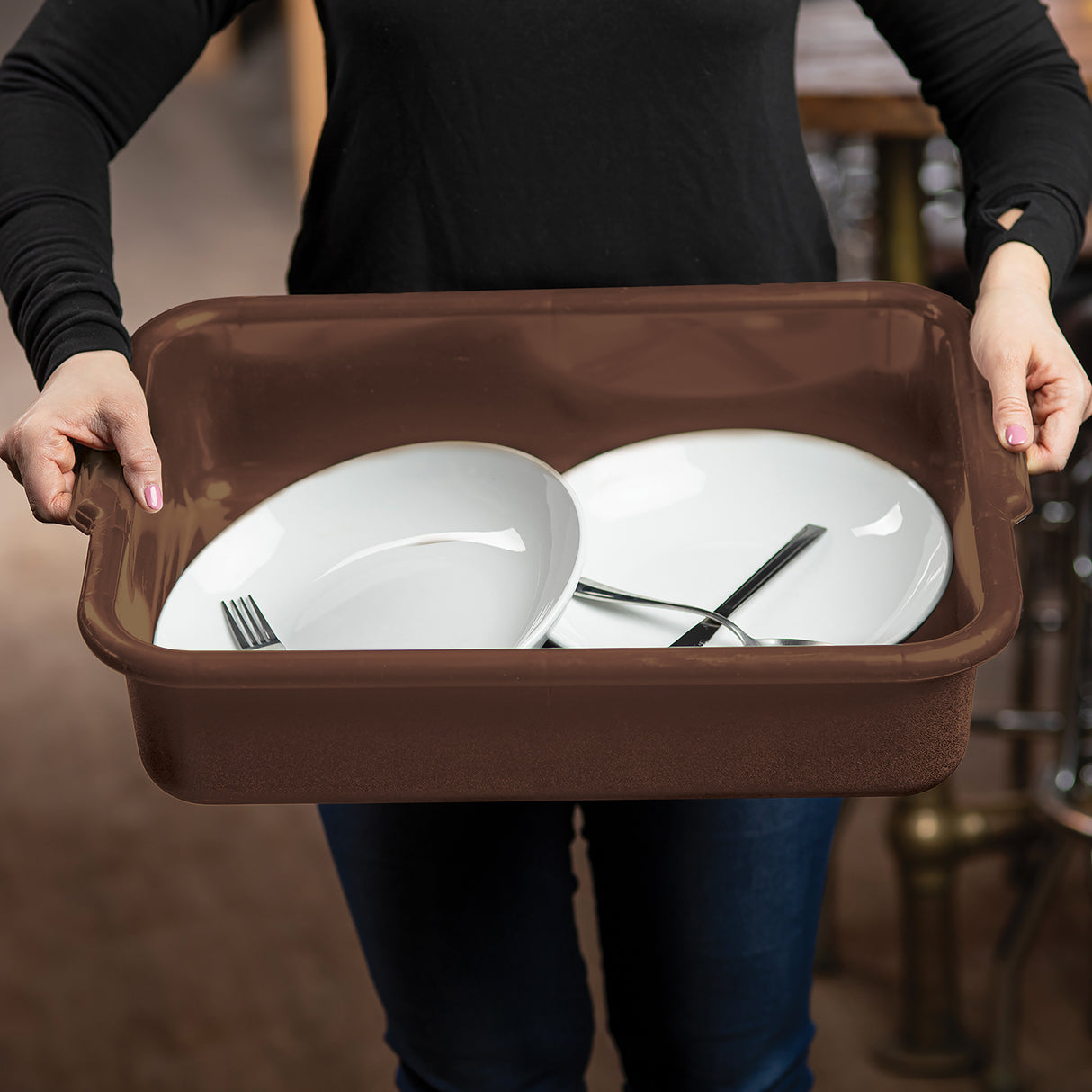 A person in a black long-sleeve shirt holds a brown stackable Tote Box containing two white plates and a fork and knife crossed over one plate, suggesting dish clearing in a restaurant or kitchen setting. The background is softly blurred.