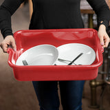 A person in a black long-sleeve shirt holds a stackable red tote box made of High Density Polyethylene, containing two white plates and some silverware. The background is blurred, with hints of a wooden table and metal pipes.
