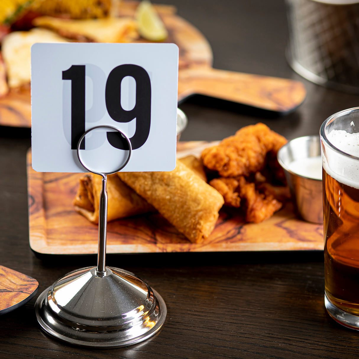 A table at a restaurant displays a TableCraft stainless steel number stand holding a card with the number 19. Behind it, there's a wooden board with fried food, including chicken tenders, an egg roll, dipping sauce, and a glass of beer.