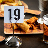 A table at a restaurant displays a TableCraft stainless steel number stand holding a card with the number 19. Behind it, there's a wooden board with fried food, including chicken tenders, an egg roll, dipping sauce, and a glass of beer.