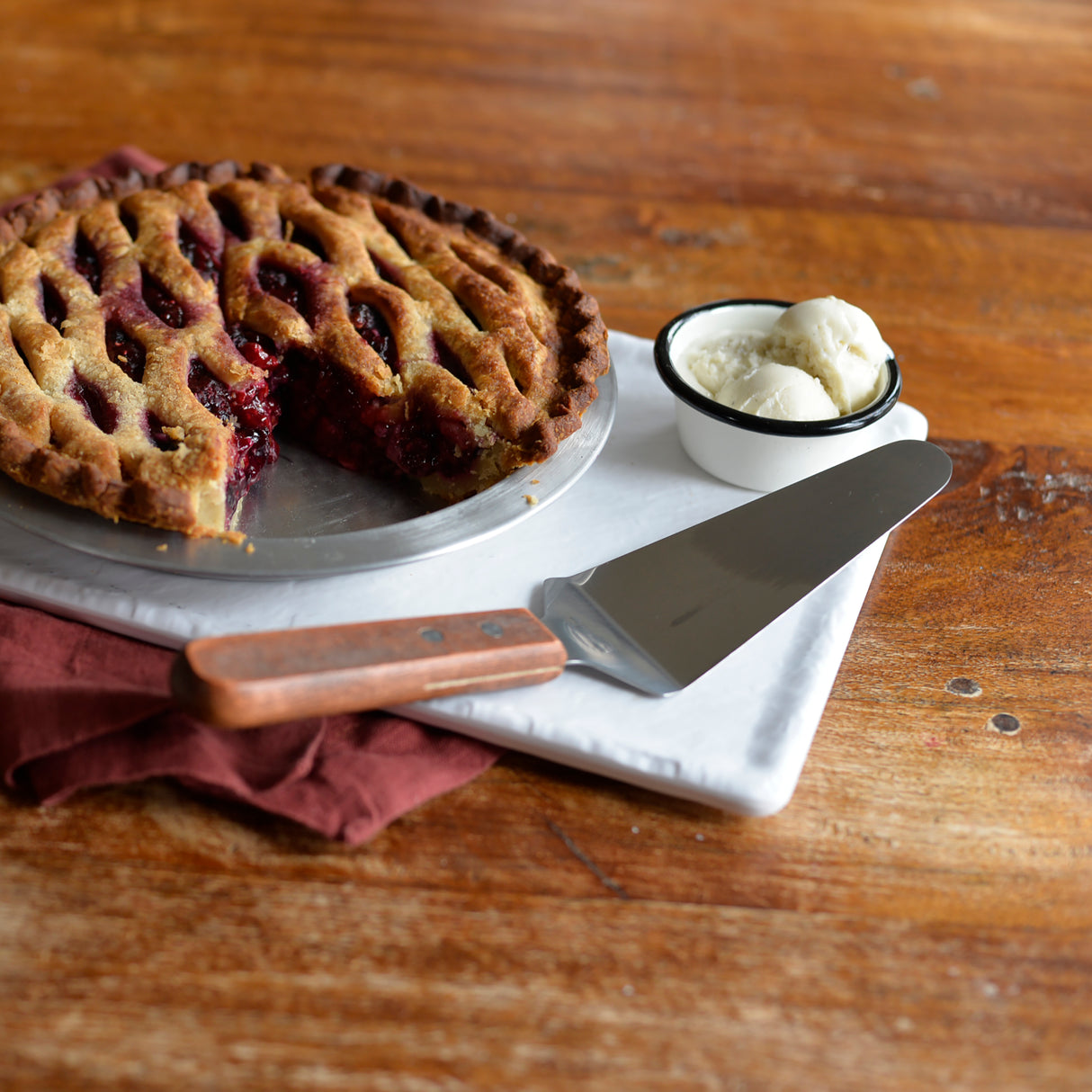 A lattice-top berry pie with a slice missing sits on a white tray beside a pie server featuring a wood handle and stainless steel blade. A small bowl of two scoops of vanilla ice cream is nearby on the rustic wooden table with a folded burgundy napkin.