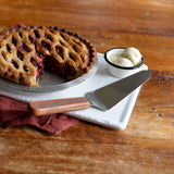 A lattice-top berry pie with a slice missing sits on a white tray beside a pie server featuring a wood handle and stainless steel blade. A small bowl of two scoops of vanilla ice cream is nearby on the rustic wooden table with a folded burgundy napkin.