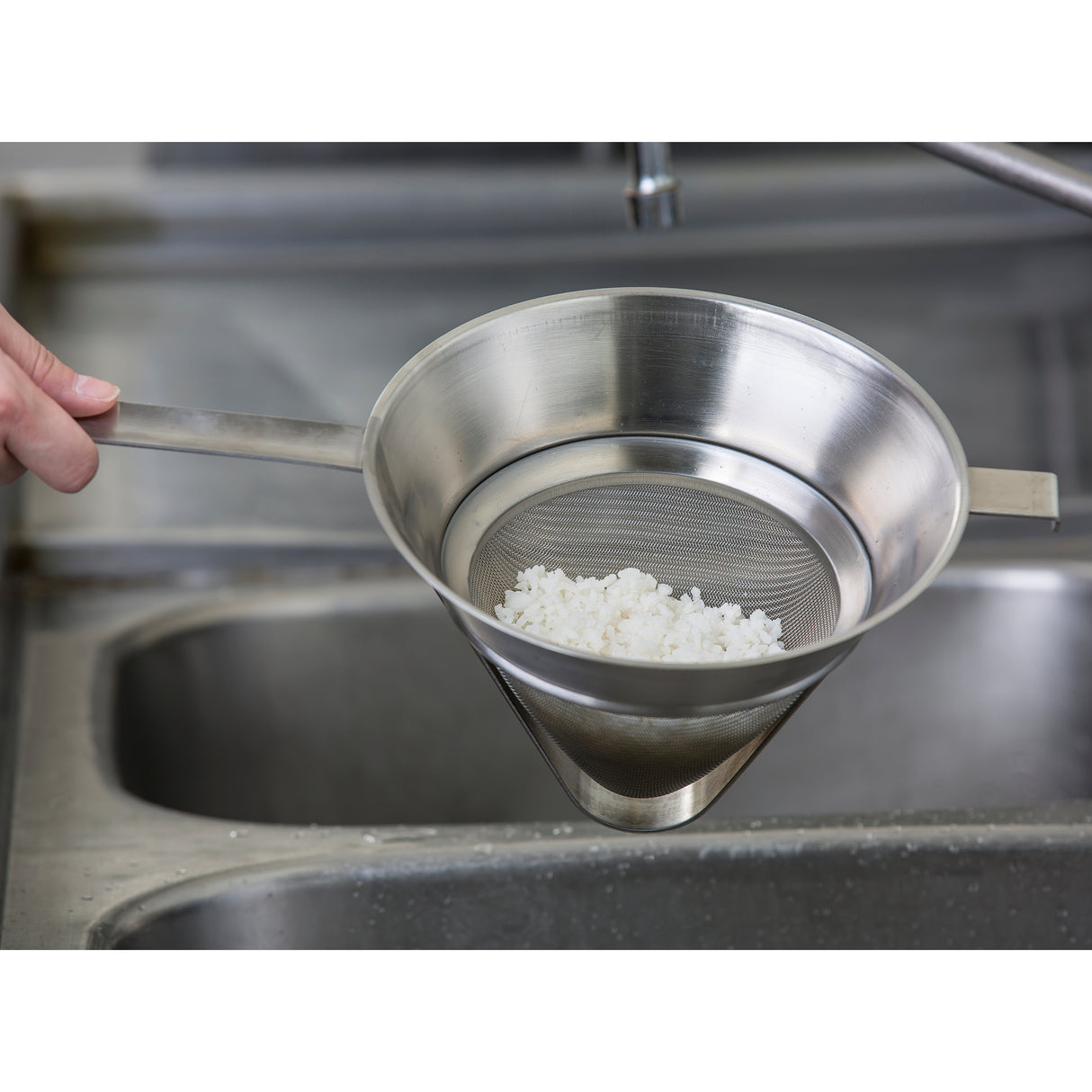 A hand holds a stainless steel Bouillon strainer with a handle over a sink. Cooked white rice sits inside the fine mesh, and water drains through. The background features part of the stainless steel sink and faucet.