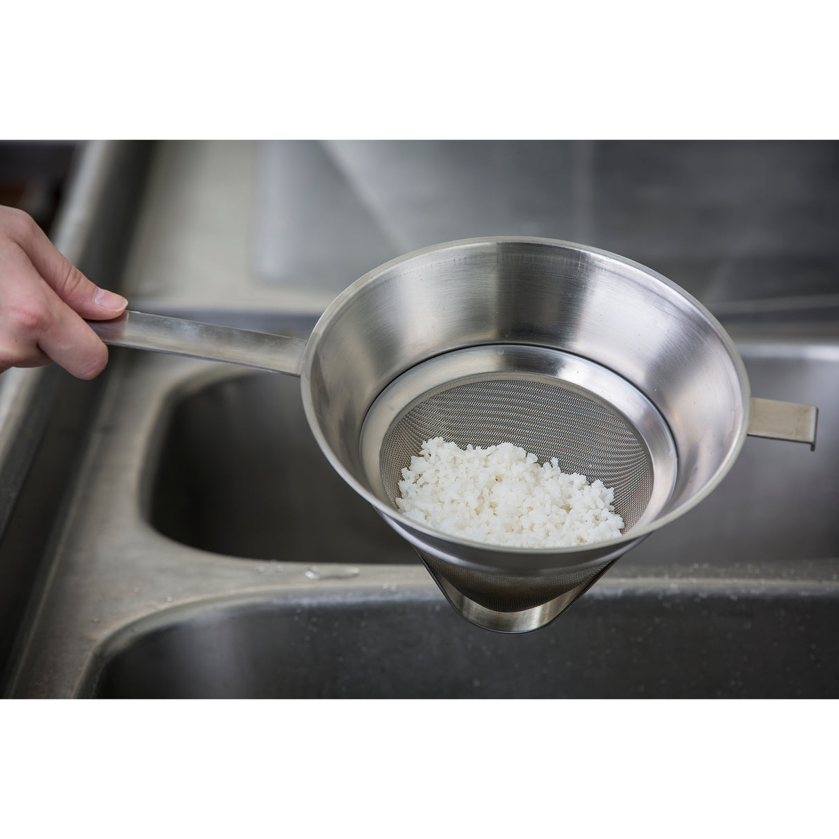A hand holds a stainless steel bouillon strainer with cooked white rice inside, draining over a kitchen sink. The background shows a metallic sink and countertop, highlighting the fine mesh strainer and rice.
