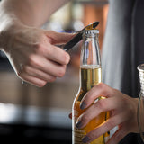 Close-up of two hands opening a glass bottle of beer with a stainless steel pocket bottle opener. The person’s fingernails are painted gray, and the light golden liquid glows inside. The softly blurred background keeps focus on the hands and bottle.
