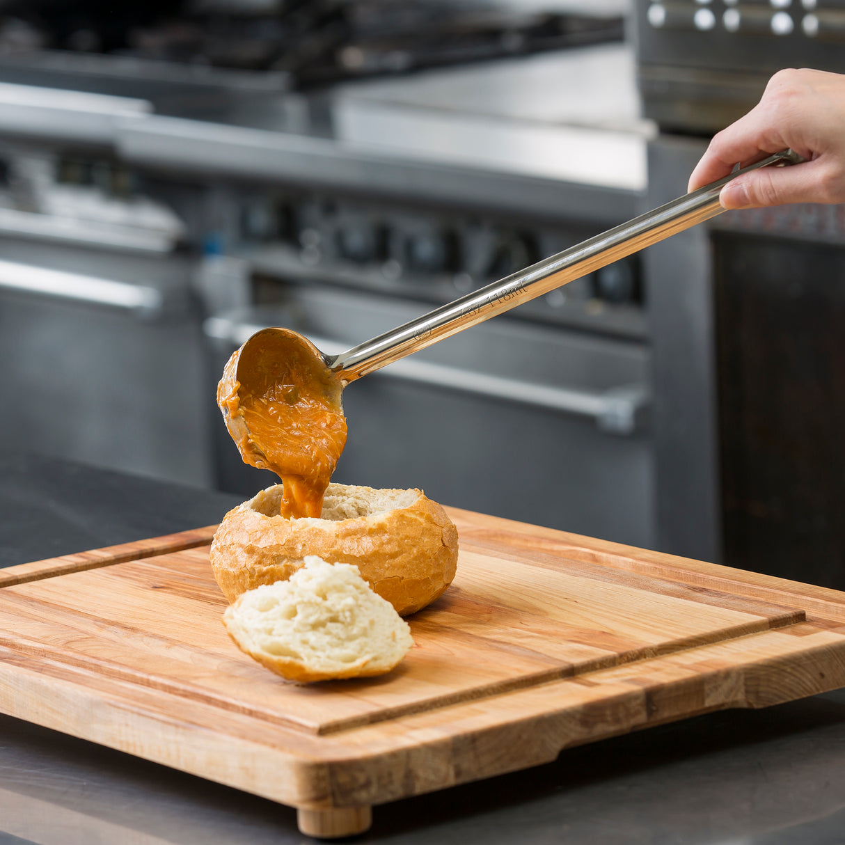 A hand pours thick, orange soup from a TableCraft stainless steel ladle into a hollowed-out bread bowl on a wooden cutting board. The bread top rests beside the bowl, with a blur of metal equipment from a commercial kitchen in the background.