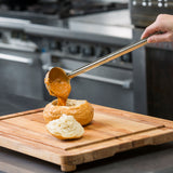 A hand pours thick, orange soup from a TableCraft stainless steel ladle into a hollowed-out bread bowl on a wooden cutting board. The bread top rests beside the bowl, with a blur of metal equipment from a commercial kitchen in the background.
