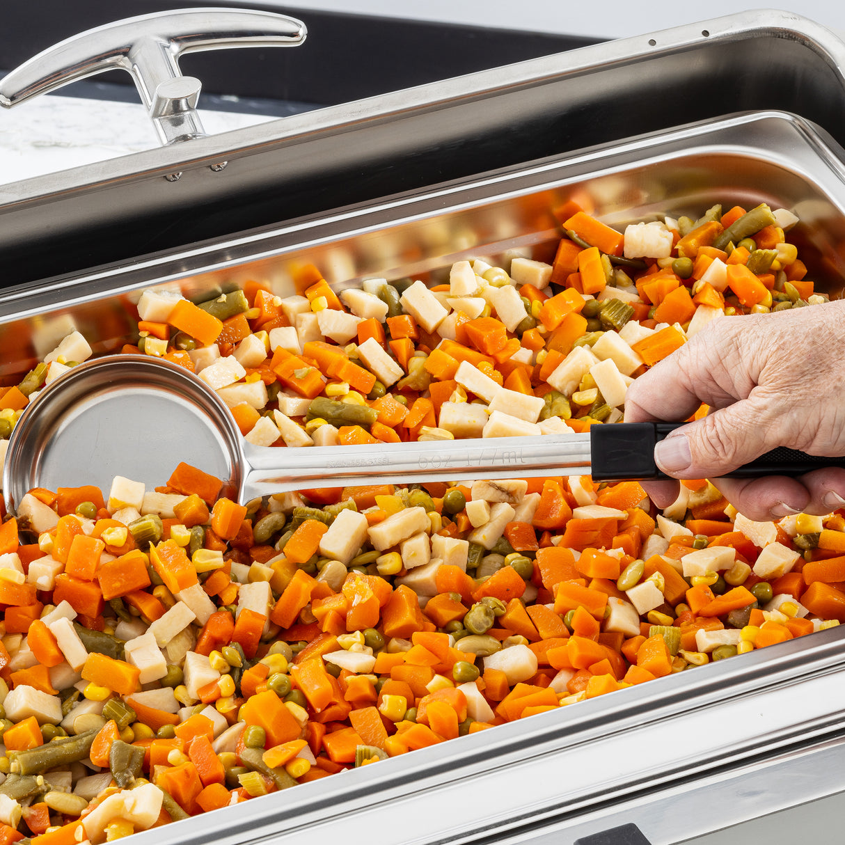 A close-up of a catering tray filled with mixed vegetables: diced carrots, green beans, corn, and parsnips. A hand holding a stainless steel serving spoon is scooping the vegetables. The tray sits on a buffet line under a shiny metal lid.