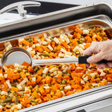 A close-up of a catering tray filled with mixed vegetables: diced carrots, green beans, corn, and parsnips. A hand holding a stainless steel serving spoon is scooping the vegetables. The tray sits on a buffet line under a shiny metal lid.