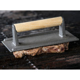 A sturdy steak weight with a wood handle presses down on cooked meat atop a dark, ridged grill. Its cast aluminum build ensures even flattening, while the blurred background keeps focus on the press and perfectly seared meat.